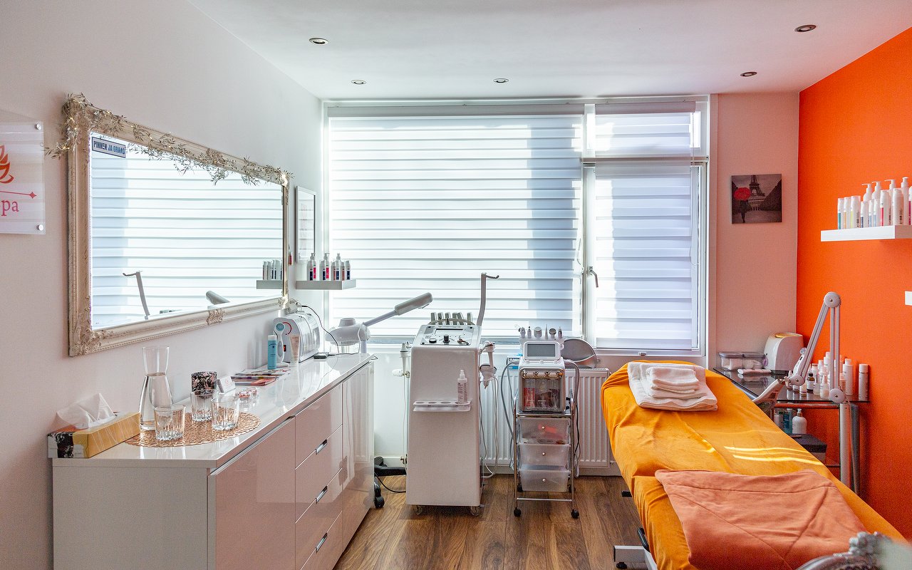 Behandelkamer met spiegel, raam, apparatuur en een behandelbank met oranje handdoeken